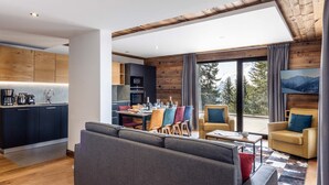 A modern living room with a sofa, dining table, and chairs. There is a kitchen area with wooden cabinets and a TV. The room has large windows with a view of trees and mountains. - Holiday Home in La Rosiere Near Ski Slopes (Montvalezan)