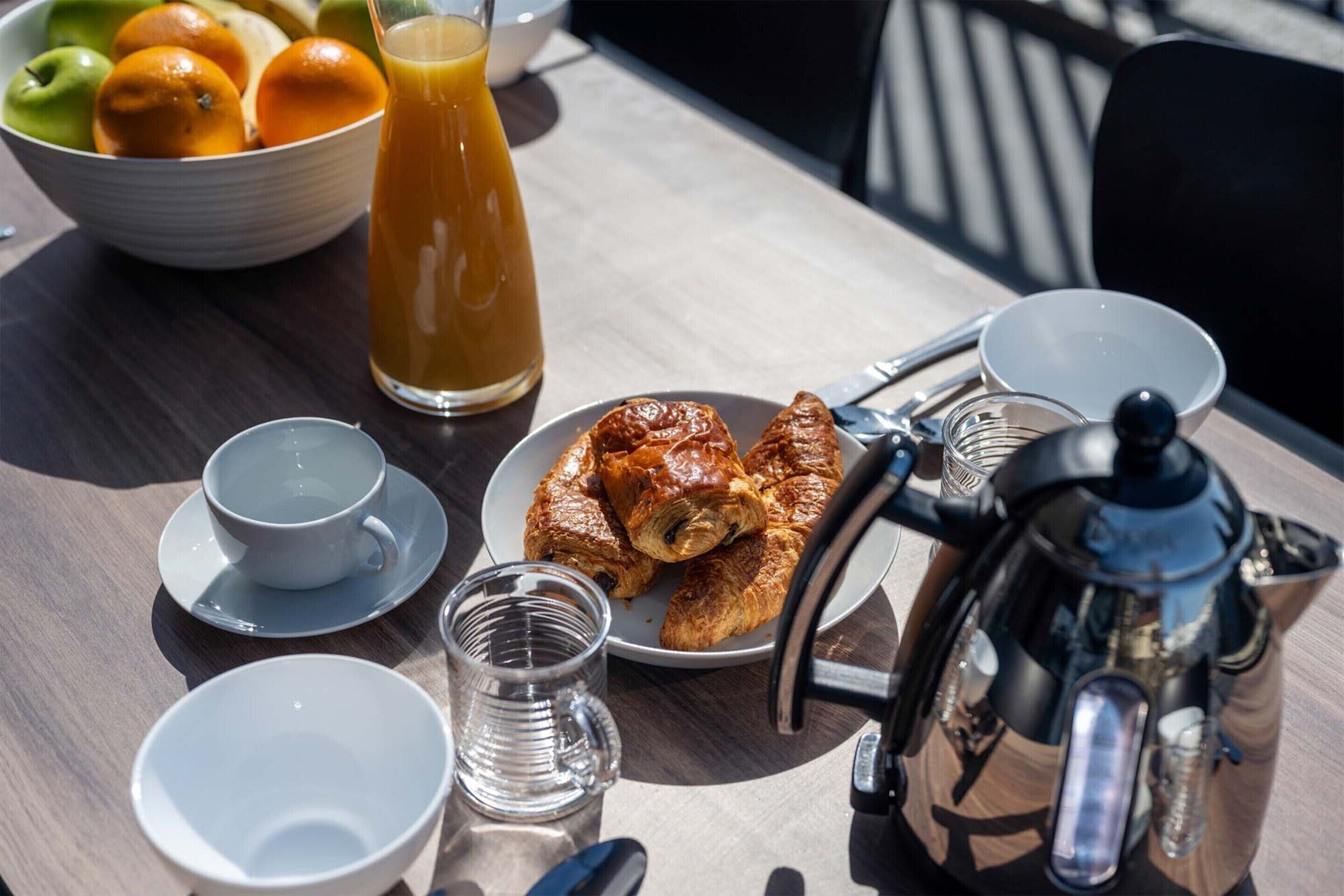 Ein Frühstückstisch mit Croissants, einer Kaffeekanne, einer Obstschale und einem Glas Orangensaft.
