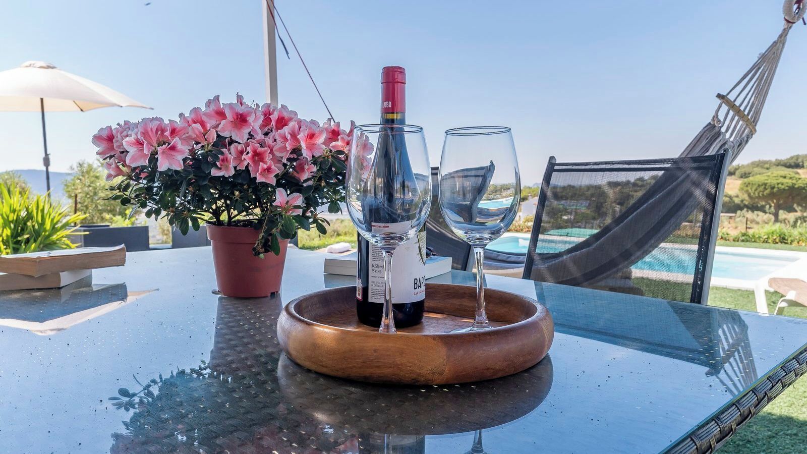 A table with a bottle of wine, two glasses, a wooden tray, and a potted plant with pink flowers.