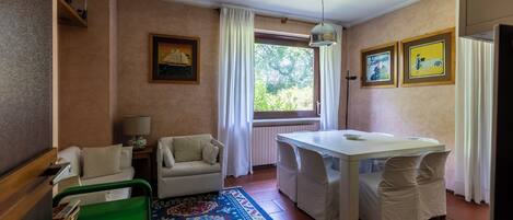 A room with a dining table, chairs, a sofa, a green chair, a patterned rug, a lamp, and framed pictures on the wall.