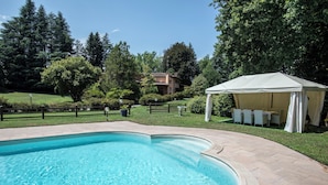A swimming pool with a curved edge, surrounded by a paved patio. There is a gazebo with a white canopy and outdoor furniture. - Villa La Fortezza - Six Bedroom Villa, Sleeps 12 (Dormelletto)