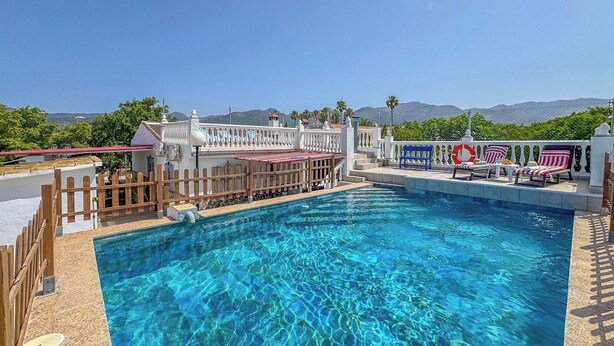 A clear blue pool with a wooden deck and lounge chairs, surrounded by a white fence and a white building with a red roof.