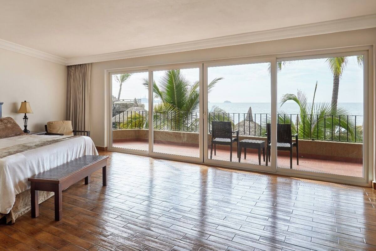 A spacious bedroom with a large bed, a bench, and a view of the ocean through large sliding doors.