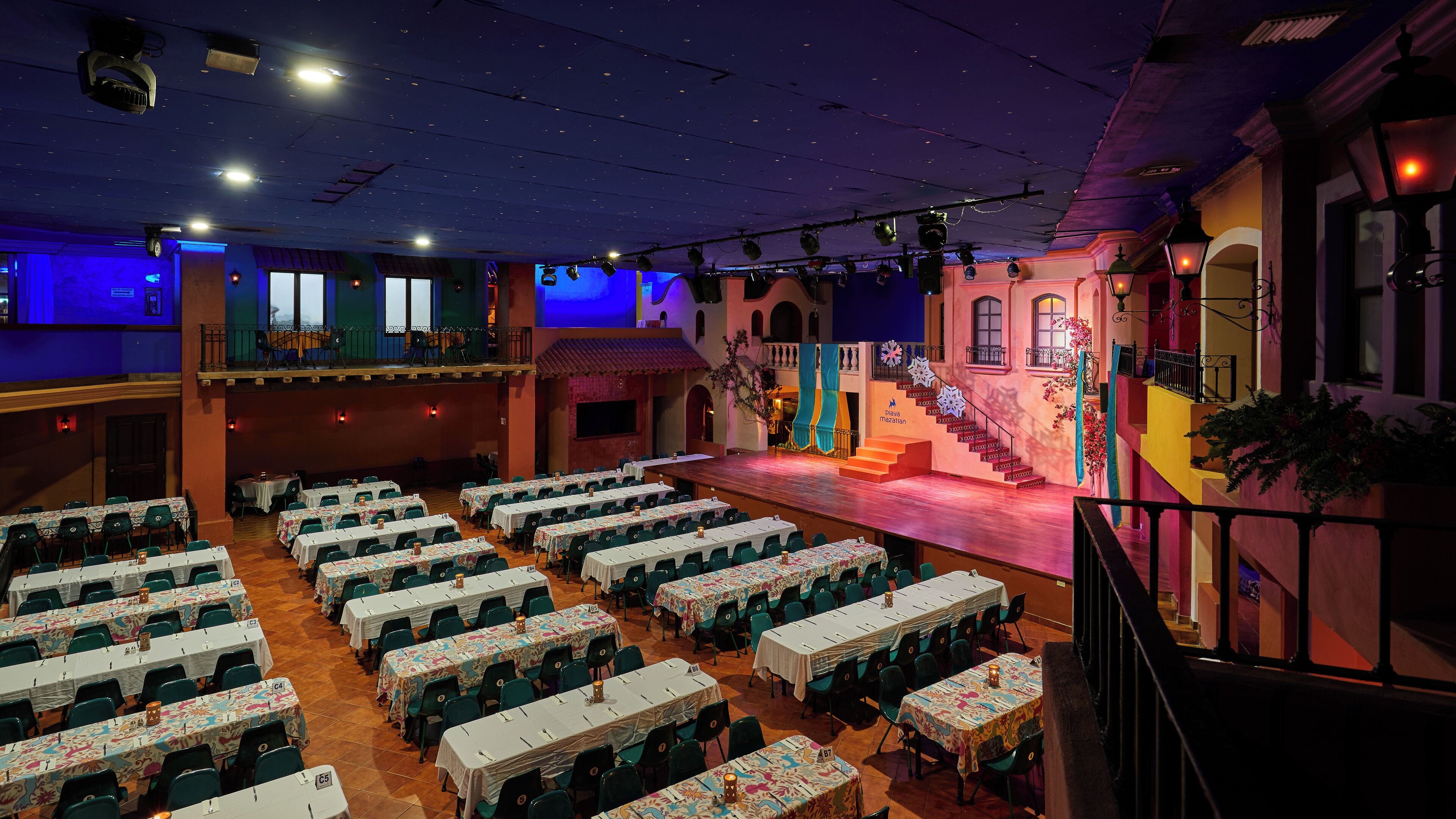A banquet hall with multiple tables and chairs, a stage with a staircase, and a balcony.