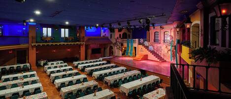 A banquet hall with multiple tables and chairs, a stage with a staircase, and a balcony.
