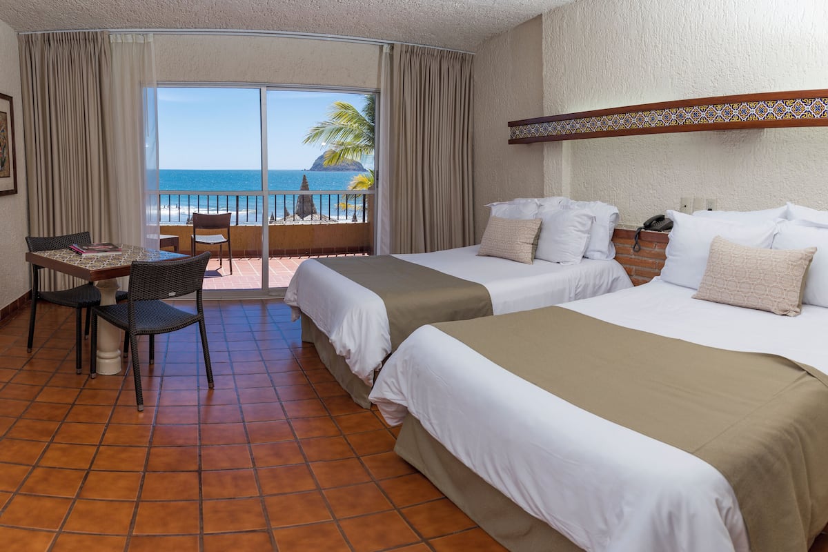 A hotel room with two beds, a table with chairs, and a balcony with a view of the ocean.