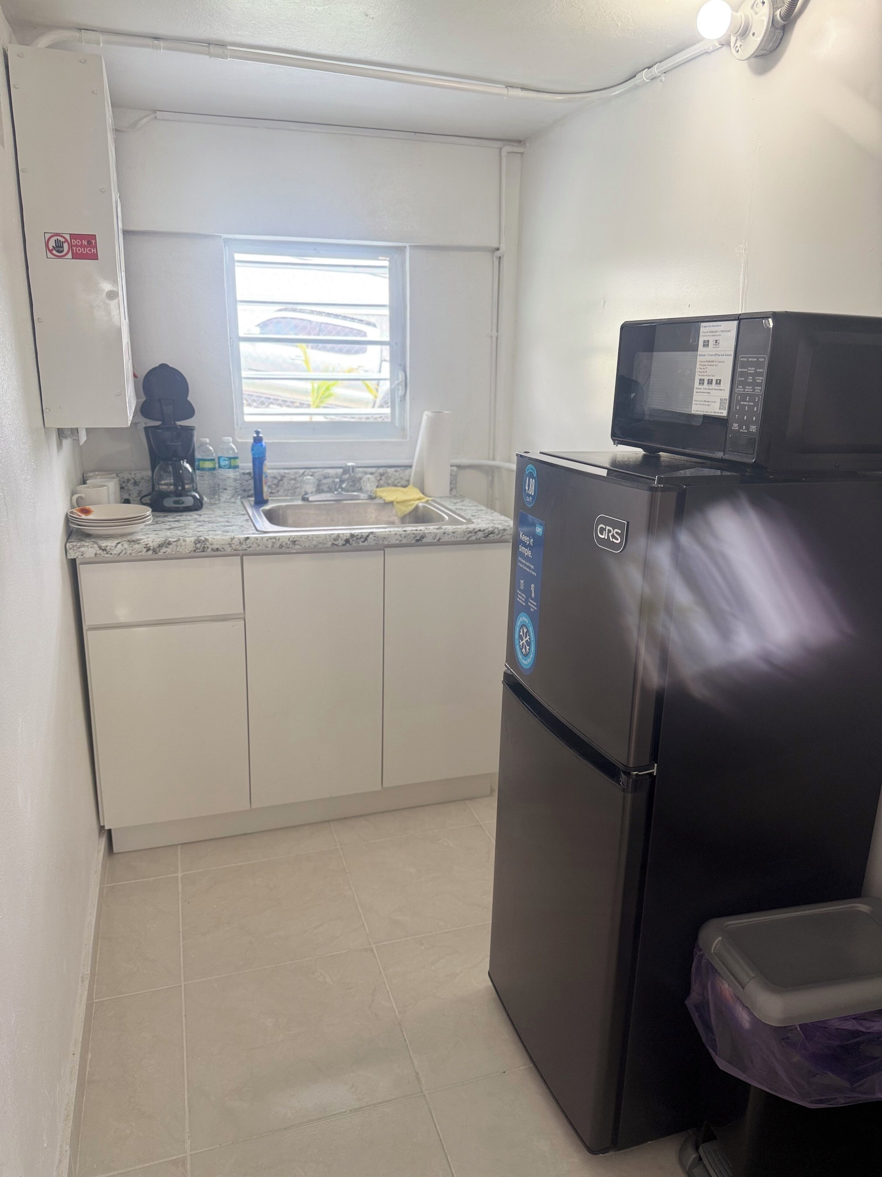 Une cuisine compacte équipée d’un réfrigérateur, d’un four à micro-ondes, d’une machine à café et d’une fenêtre avec des stores.