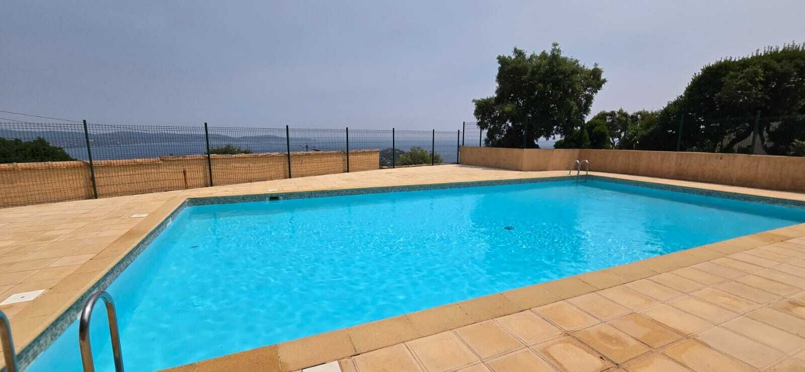 A rectangular swimming pool with a tiled deck, surrounded by a fence, and trees in the background.