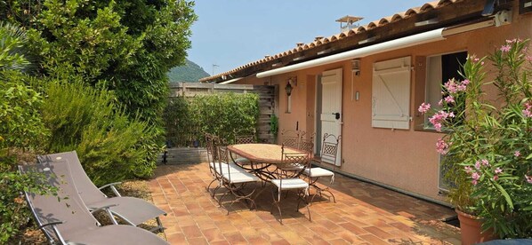 A patio with a table and chairs, surrounded by greenery and a building with a tiled roof. - Air-conditioned house with spectacular ocean views, 2 bedrooms, garden, and pool. (Cavalaire-sur-Mer)