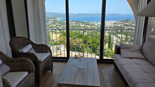 Ein Balkon mit Korbmöbeln, einem Holztisch und einer Sitzecke mit Blick auf eine Küstenstadt und das Meer.