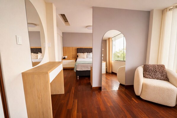 A modern hotel room with a wooden floor, a bed with a headboard, a bedside table, a chair, and a large mirror.