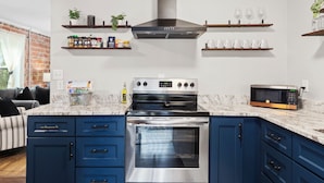 Una cocina moderna con armarios azules, horno de acero inoxidable, microondas y campana extractora. Hay estantes con utensilios de cocina y una planta en la pared.