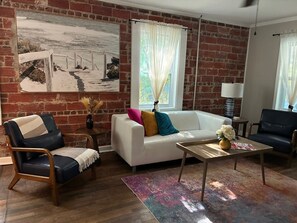 A living room with a brick wall, a white sofa, a black leather chair, a coffee table, and a painting.