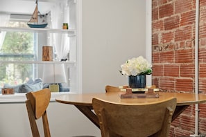 A dining area with a round table, wooden chairs, a vase with white flowers, a ship decoration, and a brick wall.
