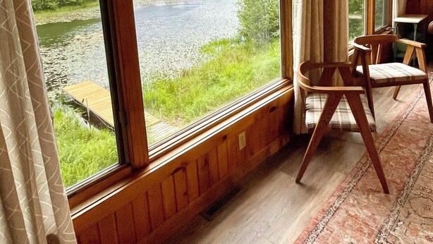 Ein Zimmer mit großen Fenstern, die auf einen Fluss und Grünflächen blicken, Holzböden und einem Sessel.