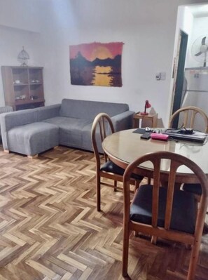 A living room with a grey sofa, wooden chairs, a herringbone-patterned floor, a painting on the wall, and a white refrigerator.
