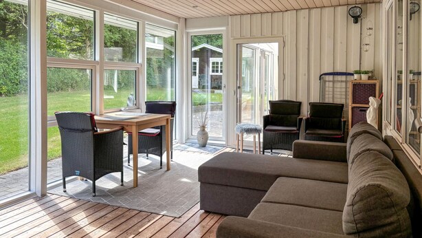 A cozy living room with a wooden floor, a grey sofa, wicker chairs, and a table. Large windows offer a view of a garden and a house.
