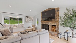 A modern living room with a stone fireplace, comfortable seating, and a large window overlooking a garden. - Beautiful and Spacious  Home with WiFi and AC in delightful Narragansett (Narragansett)