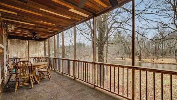 A covered outdoor area with a table and chairs, a wooden ceiling, and a view of a forest and water.