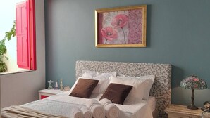Una cama bien tendida, toallas enrolladas, una lámpara de noche y una obra de arte floral enmarcada en la pared.