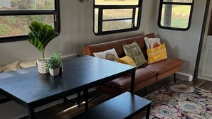 Una sala de estar acogedora con un sofá de cuero marrón, una mesa de madera oscura y un banco. Hay dos ventanas con vistas a zonas verdes y una masa de agua.