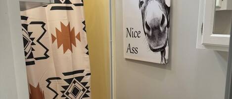 A small bathroom with a toilet, a sink, a shower curtain with a geometric pattern, a rug with a sun design, and a wall-mounted picture of a donkey with the text "Nice Ass".