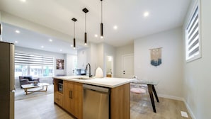 A modern kitchen with wooden cabinets, a dishwasher, and pendant lighting. There is a dining area with a glass table and chairs.