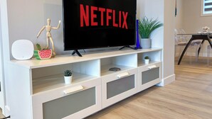 A modern living room with a flat-screen TV displaying the Netflix logo, a wooden mannequin, a watermelon-shaped speaker, and a potted plant on a white entertainment unit.