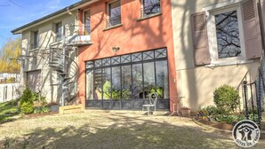 Un edificio de dos pisos con un amplio ventanal, una escalera de caracol y un pequeño jardín con plantas y un banco.