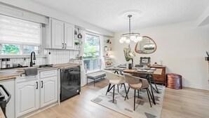 Une cuisine moderne avec des meubles blancs, un coin repas doté d’une table et de chaises en bois, et une grande fenêtre qui laisse entrer la lumière naturelle.