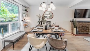 Une salle à manger moderne avec une table en bois dressée pour quatre personnes, entourée de chaises. Une grande fenêtre offre une vue sur la verdure, et on y trouve également une cheminée et un téléviseur fixé au mur.