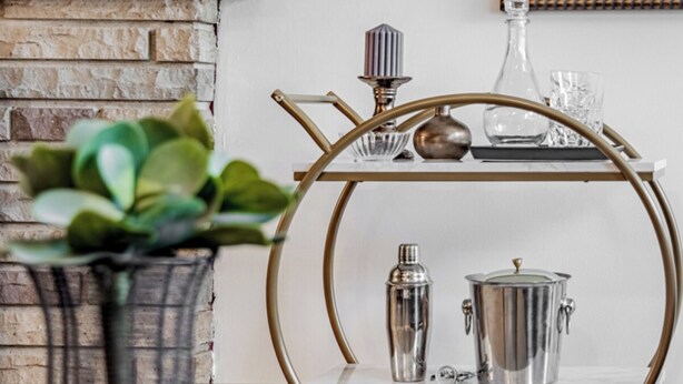 Un coin bar avec un tabouret de bar en métal, un shaker à cocktails, une bouteille et un verre posés sur une étagère.