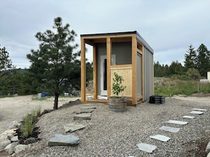 A small wooden structure with a door, a potted plant, and a gravel path leading to it. - Osoyoos Lake View | 2BR Hot Tub Retreat (Osoyoos)