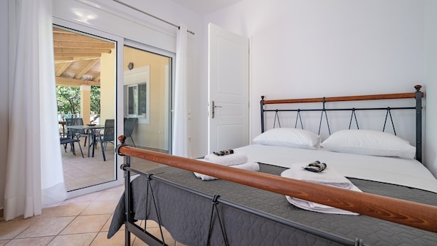 A bedroom with a bed, a wooden headboard, a view through an open door to a dining area, and a balcony with a table and chairs. - G&A Family Home near the Beach by MΡS (Nea Makri)