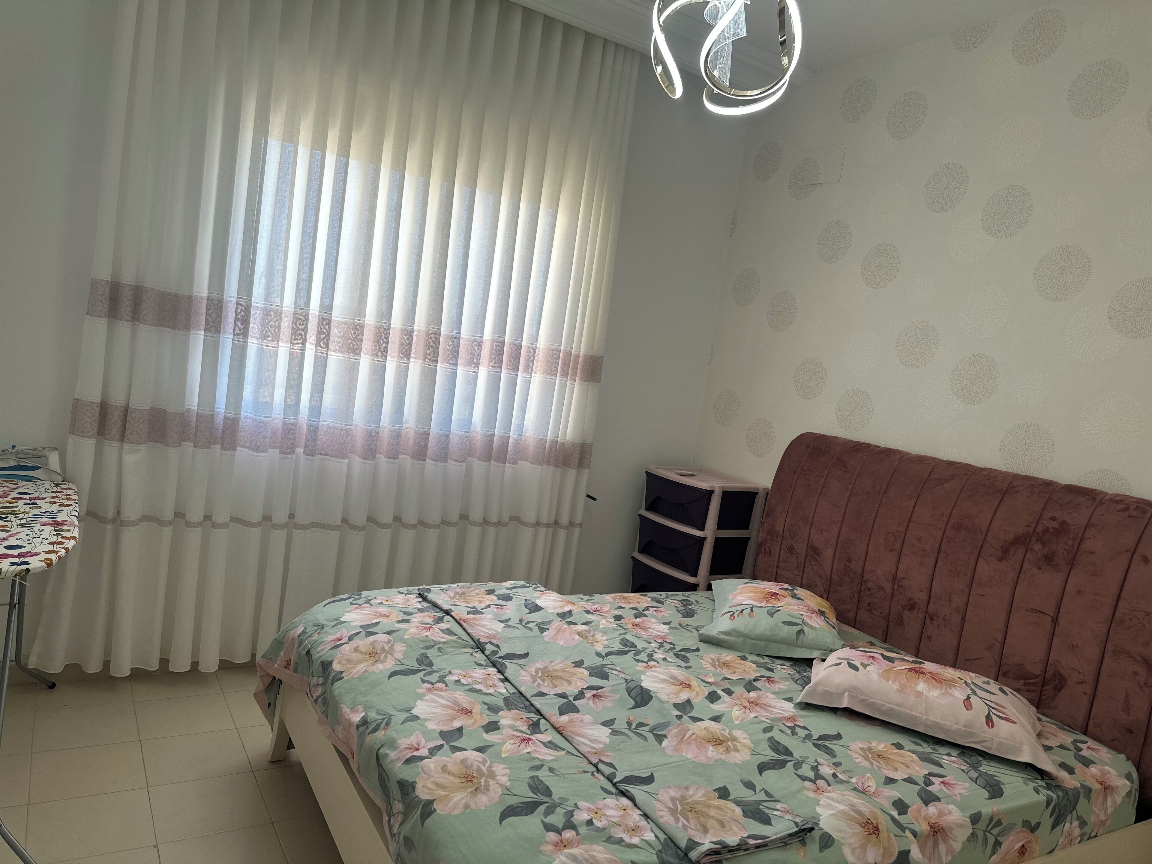 A bedroom with a bed, a bedside table, a chair, a window with curtains, and a ceiling light fixture.