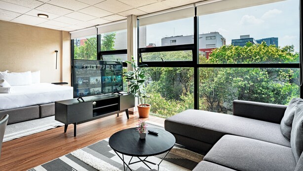 A modern hotel room with a large bed, a sofa, a TV, and a view of the city through large windows. - VH  Loft moderno en Cibeles Roma Norte  169 (Ciudad de México)