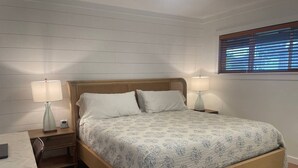Een slaapkamer met een houten bed, twee nachtkastjes met lampen, een bureau met stoel, en een raam met jaloezieën.