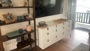 Een woonkamer met een aan de muur bevestigde televisie, een wit kastje met lades en een houten plank met diverse spullen.