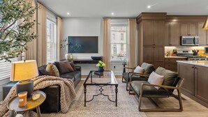 A modern living room with a sofa, coffee table, and a TV. There is a kitchen area with cabinets and appliances.