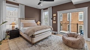 A bedroom with a bed, bedside tables, a ceiling fan, and a view of a brick building through a large window.