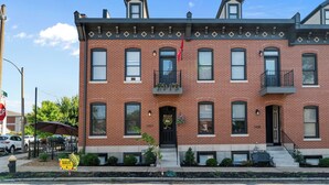 A two-story brick building with a black front door, multiple windows, and a small balcony with plants.