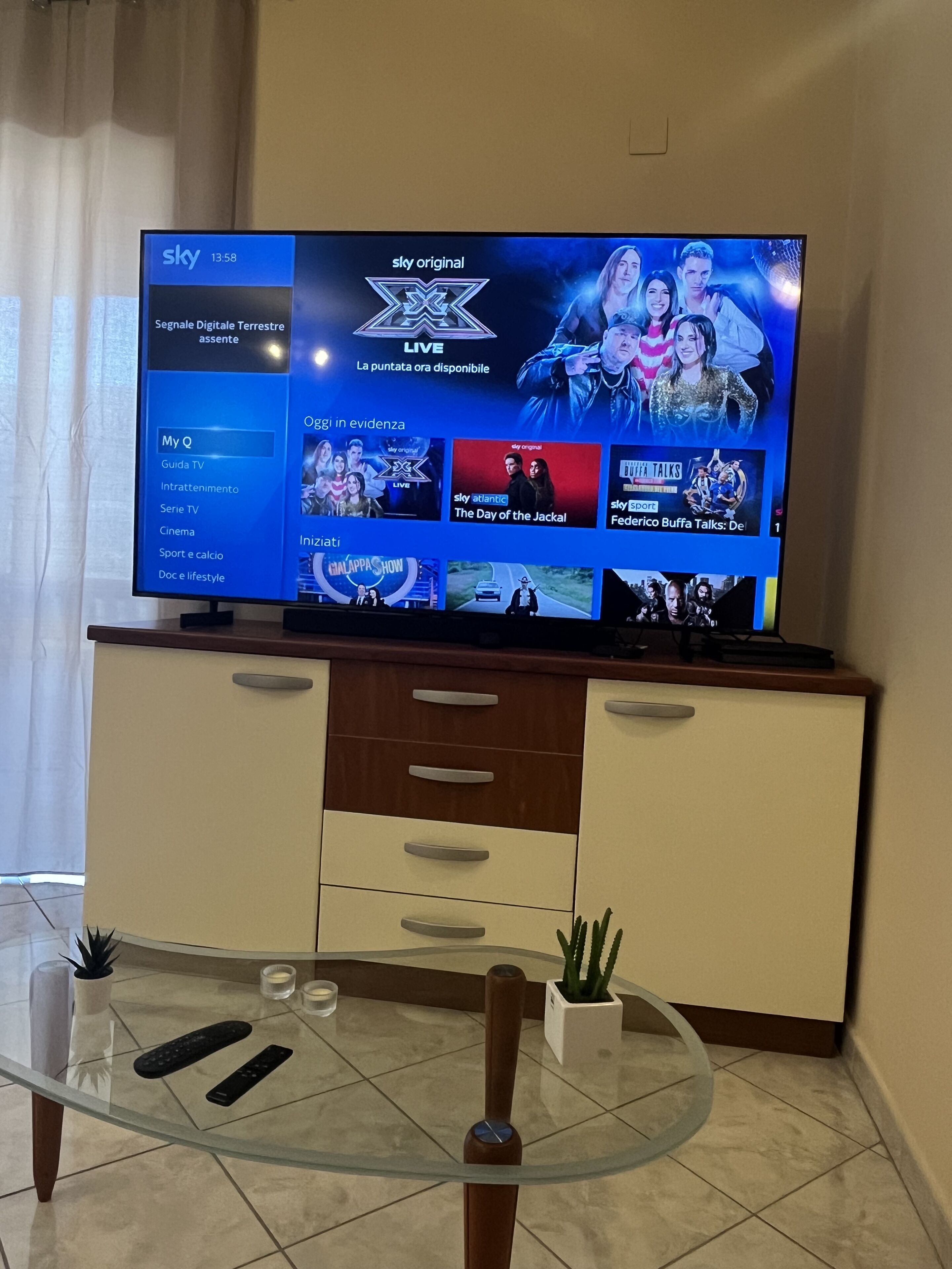 A television displaying a live show with a blue interface, a glass table with remote controls, and a wooden cabinet below.
