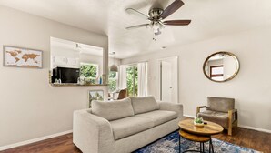 A living room with a sofa, a round coffee table, a chair, a ceiling fan, a world map on the wall, and a kitchen area visible through an open door. - Serene SoCo Escape — Walk to Cafes + Free Parking/In-Unit Laundry! (Austin)