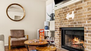 A cozy living room with a brick fireplace, a round mirror, a comfortable chair, and a small table with a board game and glasses. - Serene SoCo Escape — Walk to Cafes + Free Parking/In-Unit Laundry! (Austin)