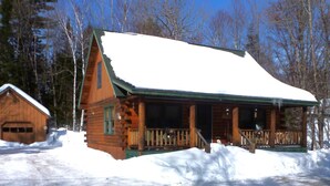 A log cabin with a porch and a covered front door, surrounded by snow and trees.