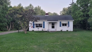 A single-story house with a dark roof, white exterior, and blue shutters. It is surrounded by a well-maintained lawn and trees.