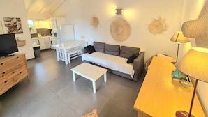 A modern living room with a sofa, a coffee table, and a TV stand. There is a kitchen area with cabinets and a refrigerator. The room has a high ceiling with exposed beams. - F2 - Casina n° 5 - 1 Bedroom - Sea View (PORTO VECCHIO)