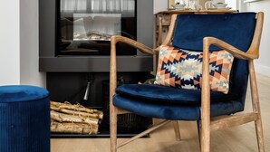 Ein modernes Wohnzimmer mit Wandverkleidung aus dunklem Holz, einem blauen Samt-Sessel mit gemustertem Kissen, einem runden Spiegel und einem Kamin mit Glastür.