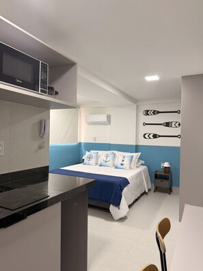 Une chambre comprenant un lit, une table de chevet, une commode, un climatiseur fixé au mur, et un coin cuisine avec un four à micro-ondes et une cuisinière.