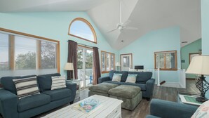 A spacious living room with a high ceiling, a ceiling fan, and large windows with blinds.